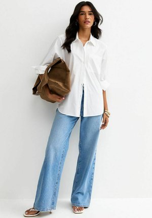 Mujer con camisa blanca abotonada, vaqueros de pierna ancha color azul claro, sandalias beige, sosteniendo un bolso grande marrón sobre un fondo blanco.