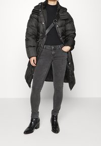 Abrigo acolchado negro con cremallera, gran cuello y tirantes en los hombros; combinado con jeans skinny gris oscuro y botas negras de tacón con un acabado texturizado.