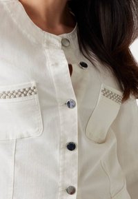 Vaalea beige takki, jossa pyöreä kaula-aukko, nepparit, kaksi etutaskua ja helmiäiset yksityiskohdat taskun reunoissa. Sileä kangaspinta.