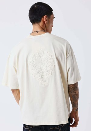 Man met kort zwart haar die een off-white oversized T-shirt draagt met een reliëf bloemenpatroon op de rug en een getatoeëerde rechterarm.