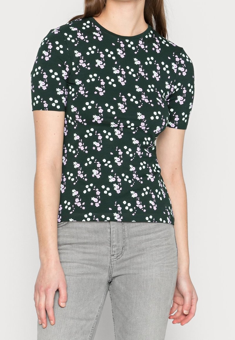Femme portant un haut à manches courtes vert foncé avec un petit imprimé floral blanc et rose, associé à un jean gris clair, debout devant un fond uni.
