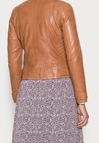 Tan leren jack met een fitted ontwerp, voorzien van textuurpanelen aan de zijkanten en naaddetails. Draag het over een kledingstuk met een bloemenpatroon.