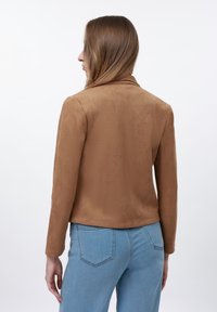 Femme aux longs cheveux bruns portant une veste en daim brun ajustée et un jean bleu clair, se tenant contre un fond blanc, de dos.