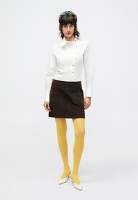 Camisa branca com botões dourados, saia castanha de camurça com bolsos frontais, collants amarelos e sapatos brancos de salto alto. Touca multicolorida.
