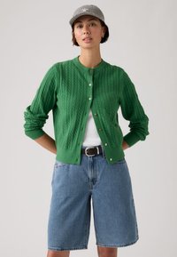 Femme aux cheveux courts portant une casquette grise à motifs, un cardigan vert en tricot torsadé sur un haut blanc, un short en jean bleu et une ceinture noire, posant avec les mains derrière le dos.