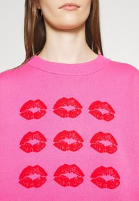 Femme portant un pull rose vif avec un motif répétitif de neuf lèvres rouges disposées en grille de trois par trois.