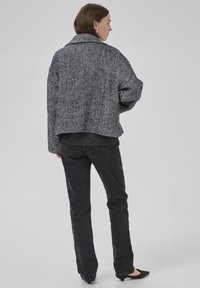 �Ženska s ramensko dolgimi rjavimi lasmi, oblečena v črno-belo herringbone jakno, temno sivo jeans hlače in črne špicate, obrnjena desno.