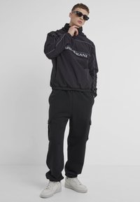 Black pullover hoodie with front zipper, elastic waist, and white logo. Paired with black cargo pants and white sneakers.