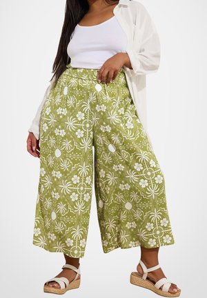 Kvinde iført grønne bukser med vide ben, dekoreret med hvide blomster- og solmotiver, hvid undertrøje, let åben skjorte og hvide kilehælssandaler.
