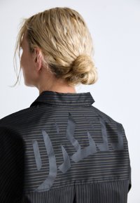 Black shirt with vertical gold stripes, featuring a large graphic on the back. The collar is flat and the fabric appears smooth and glossy.