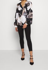 Femme portant un blouse à fleurs noires et blanches enveloppante, un pantalon noir ajusté, et des talons à bride de cheville noirs, se tenant devant un fond clair.