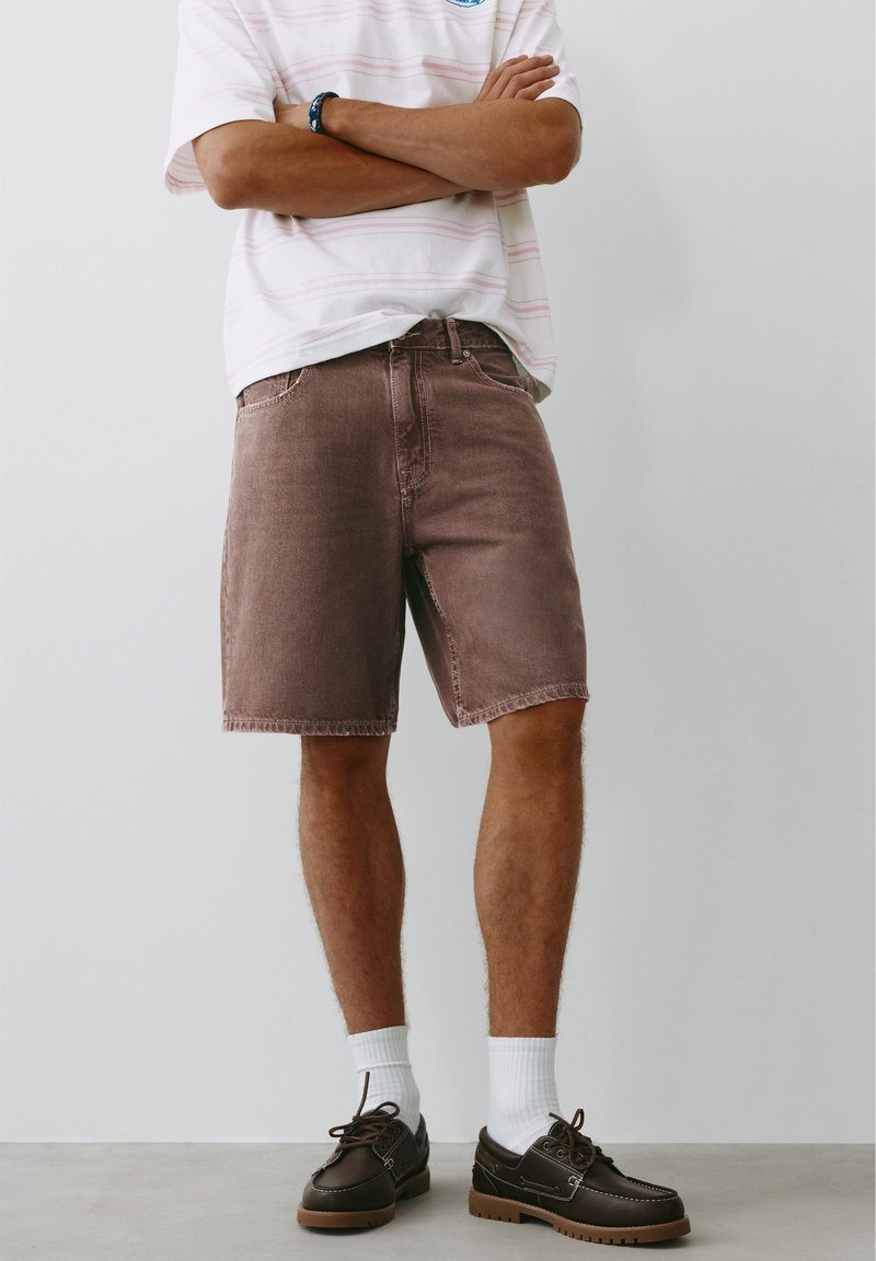 Homme debout, les bras croisés, portant un t-shirt rayé clair, un short marron jusqu'aux genoux, des chaussettes blanches et des chaussures marron foncé à lacets.