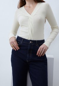 Cremfarbener, gerippter Langarmoberteil mit V-Ausschnitt und gedrehtem Frontdetail, kombiniert mit dunkelblauen, hochgeschnittenen Jeans, die beide eng anliegen und strukturiert sind.
