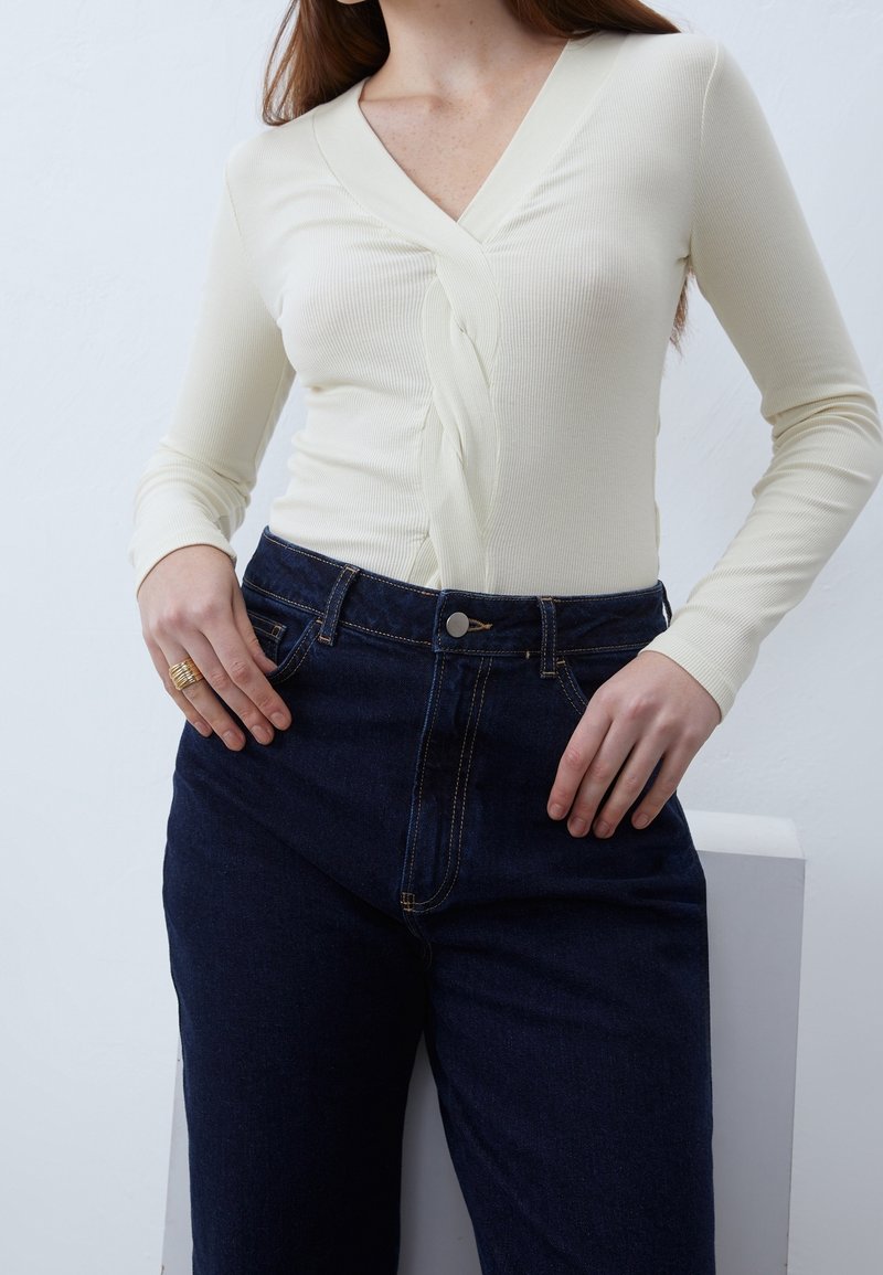 Cremfarbener, gerippter Langarmoberteil mit V-Ausschnitt und gedrehtem Frontdetail, kombiniert mit dunkelblauen, hochgeschnittenen Jeans, die beide eng anliegen und strukturiert sind.