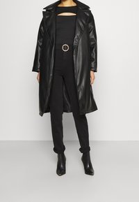 Abrigo de cuero negro con cuello de chal, de frente abierto, combinado con una blusa negra ajustada con un diseño de corte, pantalones negros y botas hasta el tobillo.