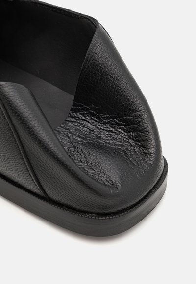 Zapato negro de cuero sin cordones con textura suave, punta redondeada y diseño de espalda abierta. Presenta una suela duradera y un detalle de costura sutil.