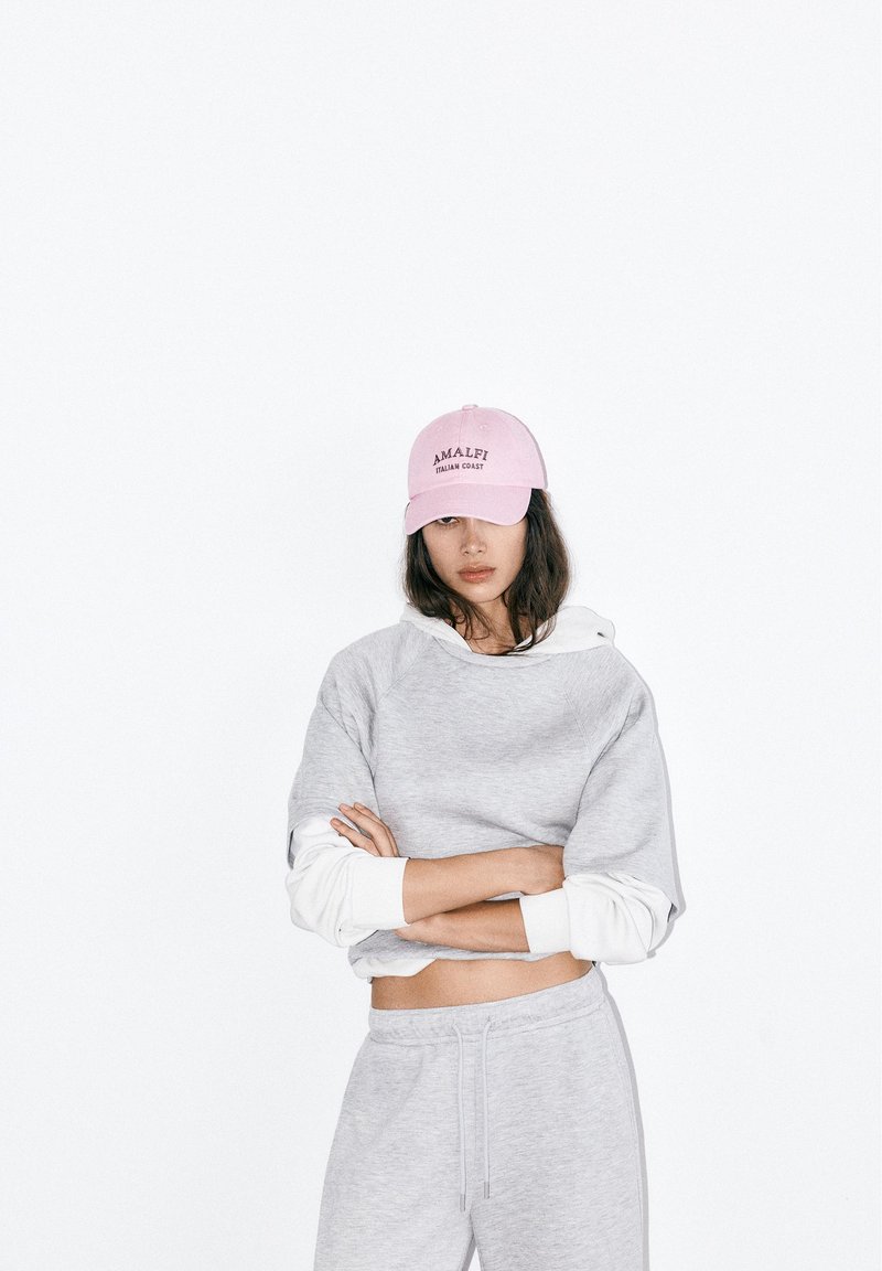 Femme portant un sweat-shirt court gris clair et un pantalon de jogging avec des manches blanches superposées en dessous, une casquette rose avec l'inscription « Amalfi Italian Coast », bras croisés.