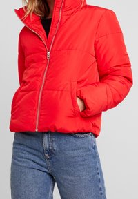 Red insulated puffer jacket with a high collar, zippered front, and side pockets. Features stitched panels for added texture.