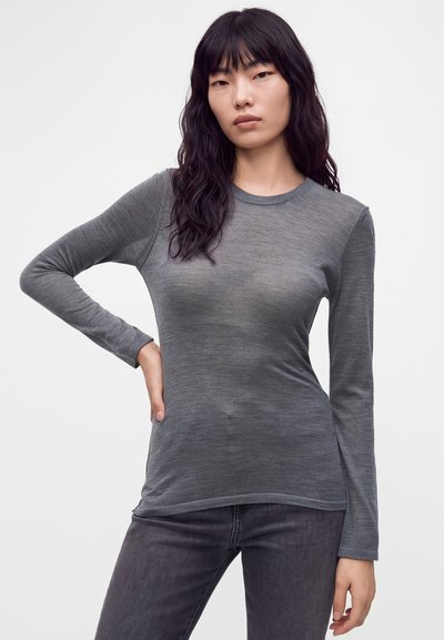 Femme aux cheveux noirs ondulés portant un t-shirt gris ajusté à manches longues et un jean gris foncé, debout avec une main sur la hanche.