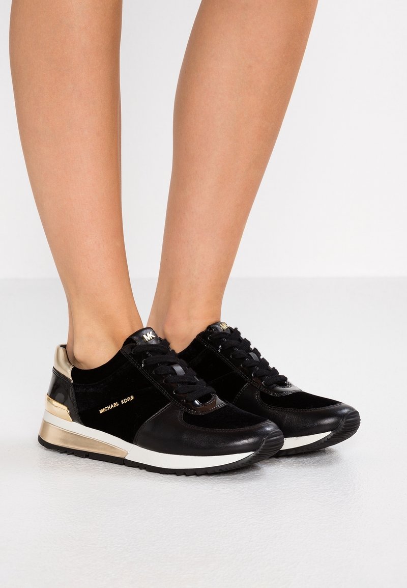Jambes portant des baskets Michael Kors noires et dorées avec des semelles intermédiaires blanches, debout sur un sol lisse gris clair.