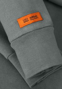Grijze sweatshirtmouw met een oranje rechthoekig stoffen label erop gestikt, met de tekst "Spitzbub Studios" en Duitse coördinaten.