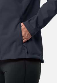 Jachetă softshell de culoare bleumarin închis, cu un buzunar cu fermoar pe lateral, textură netedă și design ajustat, purtată cu colanți negri.