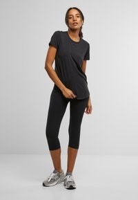 Crna majica kratkih rukava s okruglim izrezom, u kombinaciji s crnim leggingama do članaka i sivim tenisicama, na modelu protiv jednostavne pozadine.