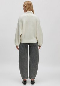 Weißer gestrickter Pullover mit hohem Kragen, oversized Passform, gerippter Struktur, kombiniert mit lockeren, dunkelgrauen Jeans und schwarzen Schuhen.