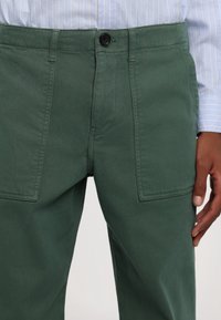 Pantalon cargo en coton vert avec de grandes poches avant, une fermeture par bouton et une finition lisse et texturée. Associé à une chemise rayée bleu clair.