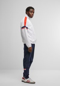 White lightweight jacket with red and navy stripes, paired with navy trousers and white sneakers with red accents. Smooth texture and relaxed fit.
