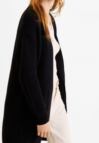 Gilet en tricot noir avec un motif texturé, design à l'avant ouvert et manches longues, porté par-dessus un haut clair et un pantalon crème.
