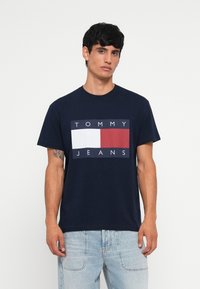 Marineblå bomulds t-shirt med et rektangulært logo i hvide, røde og blå sektioner. Korte ærmer og rund halsudskæring. Bæres med lyse jeans.