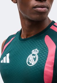 Gros plan d’une personne portant un maillot de sport vert foncé avec des accents roses et les logos du Real Madrid et d’Adidas sur la poitrine.