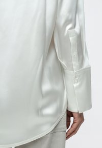 Prim-plan al unei persoane care poartă o cămașă albă din satin cu mâneci lungi și manșete cu nasturi și pantaloni bej, cu mâna odihnindu-se pe lângă corp.