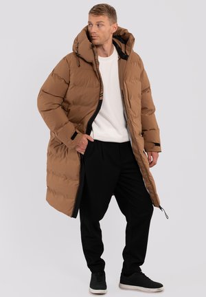 Abrigo acolchado marrón con capucha, con cierre de cremallera y textura acolchada. Lleva puesto sobre una camisa blanca y pantalones negros, complementado con zapatos negros.