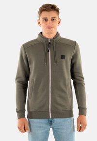Giovane uomo che indossa una giacca olive verde con zip e cordini e jeans azzurri, in piedi davanti a uno sfondo bianco semplice.