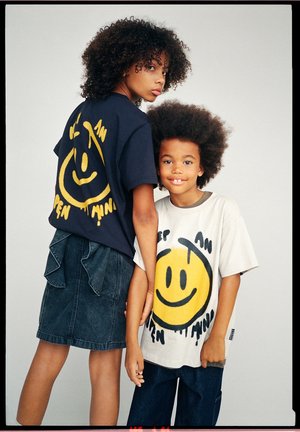 Deux enfants aux cheveux bouclés portent des t-shirts oversize avec de grands visages jaunes souriants et se tiennent proches en se tenant la main.