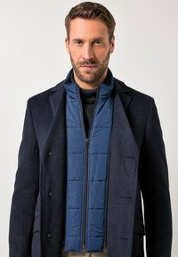 Granatowa pikowana kamizelka z wysokim kołnierzem, zapinana na zamek, z pikowanym wzorem, noszona pod ciemnym, teksturowanym płaszczem. Neutralne tło.