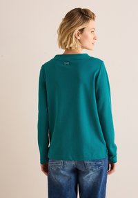 Turquoise lange mouw shirt met een kraag, voorzien van een klein label op de achterkant en zijslits, gecombineerd met lichtblauwe jeans.