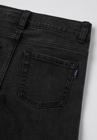 Musta farkkuhousut, joissa on teksturoitu pinta, varustettu takataskulla ja pienellä sinisellä tarralla ommeltuna vyötärölle.