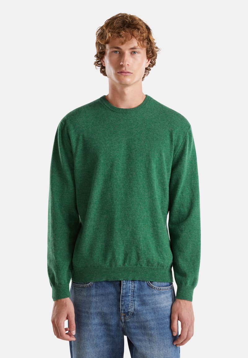 Mladý muž s kudrnatými vlasy, oblečený v zeleném svetru s kulatým výstřihem a modrých džínách, stojící na pozadí v jemně světlé barvě.