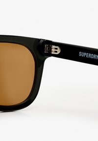 Gafas de sol negras con lentes marrones. Presentan una forma cuadrada, acabado brillante y un acento plateado en la montura marcado con "SUPERDRY."