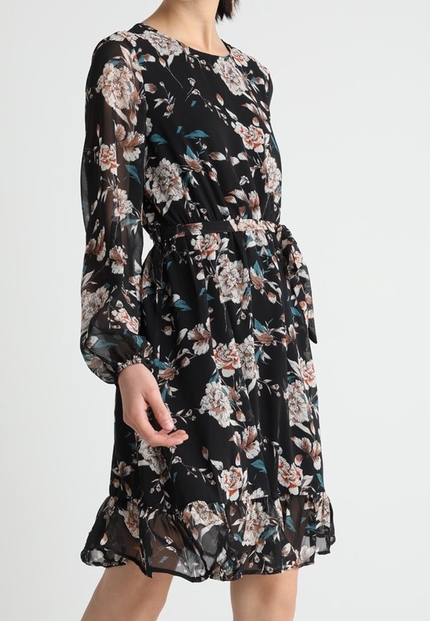 Robe florale noire avec de longues manches transparentes, taille froncée avec une ceinture et ourlet volanté portée par une personne debout.