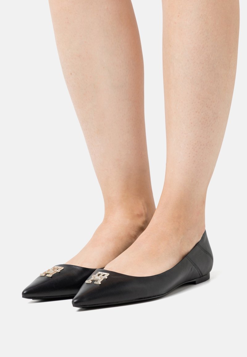 Jambes portant des ballerines pointues en cuir noir avec un logo décoratif doré sur les orteils sur un fond clair uni.