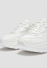 Bílé tenisky s hladkým koženým vnější povrchem, texturovanou gumovou podrážkou a tradičním šněrovacím designem. Obsahují jemné švy a robustní siluetu.