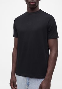 Άνδρας που φοράει μαύρο κοντομάνικο t-shirt με μικρό κεντητό κείμενο στο στήθος συνδυασμένο με ανοιχτό μπλε τζιν, εμφανιζόμενος από τους ώμους έως τους γοφούς.