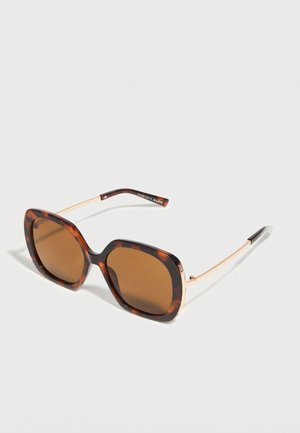 Übergroße quadratische Sonnenbrille mit Schildpattgestell, braunen Gläsern und dünnen goldenen Metallbügeln, die auf einer weißen Oberfläche liegt.