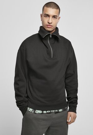 Zwarte pullover sweatshirt met een hoge kraag en een rits aan de voorkant, voorzien van een gestructureerde stof en afgeknipte mouwen, gelaagd over een gemusterd shirt.