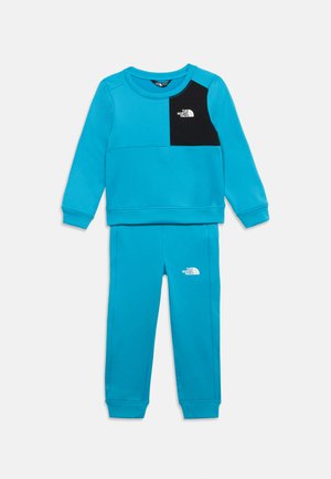 Mėlynas dviejų dalių vaikų sportinis kostiumas su ilgomis rankovėmis, ant krūtinės juodas lopas viršuje ir balti The North Face logotipai ant krūtinės ir kelnių.