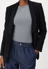 Svart kavaj över en randig, figursydd topp med en liten logotyp. Lösa denimbyxor med resår i midjan och knappdetalj fram.